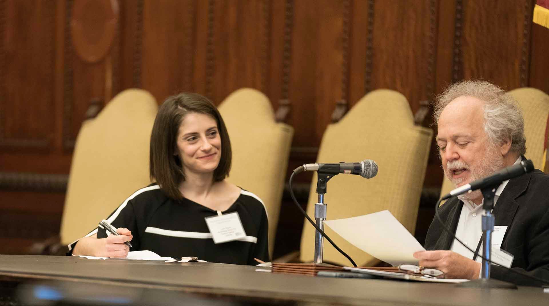 Two people sitting behind a table with ornate wood panelling behind them. One of them is speaking into a microphone while the other looks on smiling