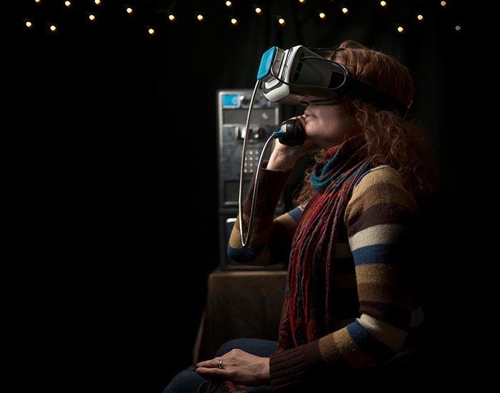 Person seated in a dark room with a VR headset on, listening to the receiver of a payphone which sits behind them